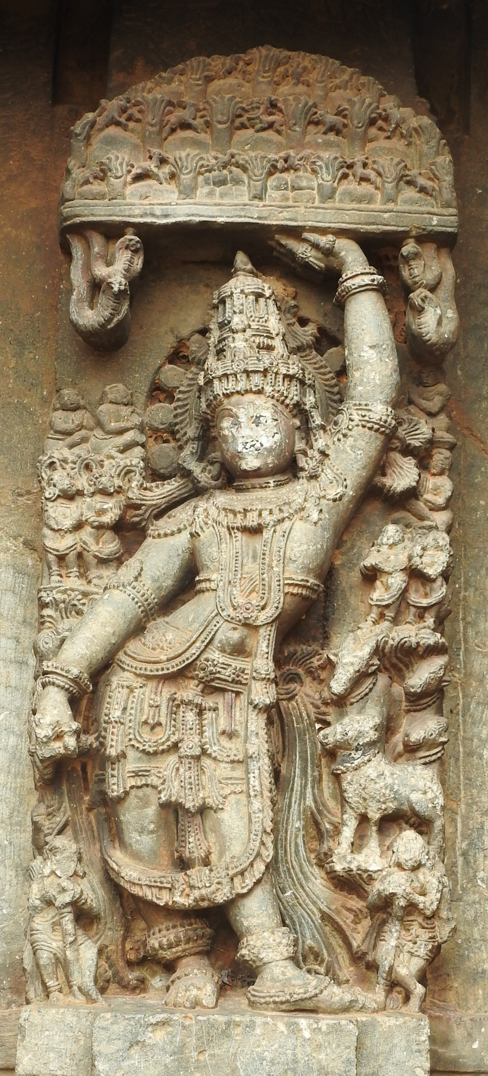 Fig. 13: The panel showing Govardhanadhari Krishna on the jangha. He is lifting Govardhana mountain aloft with his left hand and is well-ornamented. The parikara of the image displays Krishna surrounded by villagers.