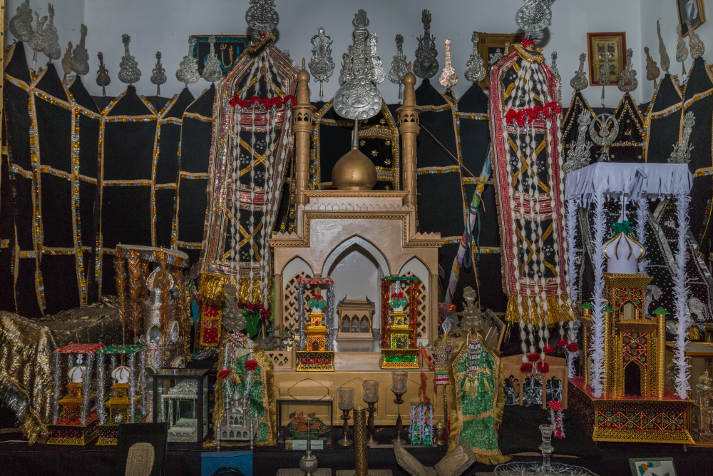 Fig. 8: A replicated rauza of Imam Hussain (a.s.) made of wood placed in an azakhana. Taziya and zarih of different sizes can be seen along with alam in the background  