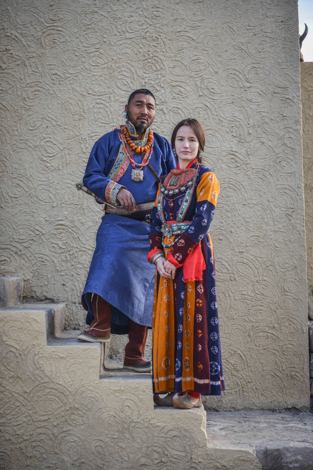 A campaign shoot for the label ‘Jigmat Couture’ shows two models dressed in contemporary yet traditional Ladakhi dress and jewellery. The man is wearing three necklaces – one amber, one with five dzi strung with coral stones, and a silver filigree worked amulet (gau) hung from a silk fabric. He wears a silver and turquoise earring, supported by a loop around his ear. A leather belt decorated with silver worked pieces placed at intervals; the dagger inserted into it has a silver hilt with a single turquoise stone. The woman is wearing a large gorget, a stiff collar piece (skebug), made from a piece of padded red cloth onto which a central silver and gold-plated amulet is stitched, surrounded by lines of coral, with a fringe of seven silver amulets. She is wearing turquoise and silver earrings. Around her waist is a turquoise and coral belt. On her right hand she is wearing a silver bracelet embossed with Buddhist and geometric patterns.   (Photographer: Bablu and Sonny Singh, 2017; Courtesy: Jigmat Couture.)