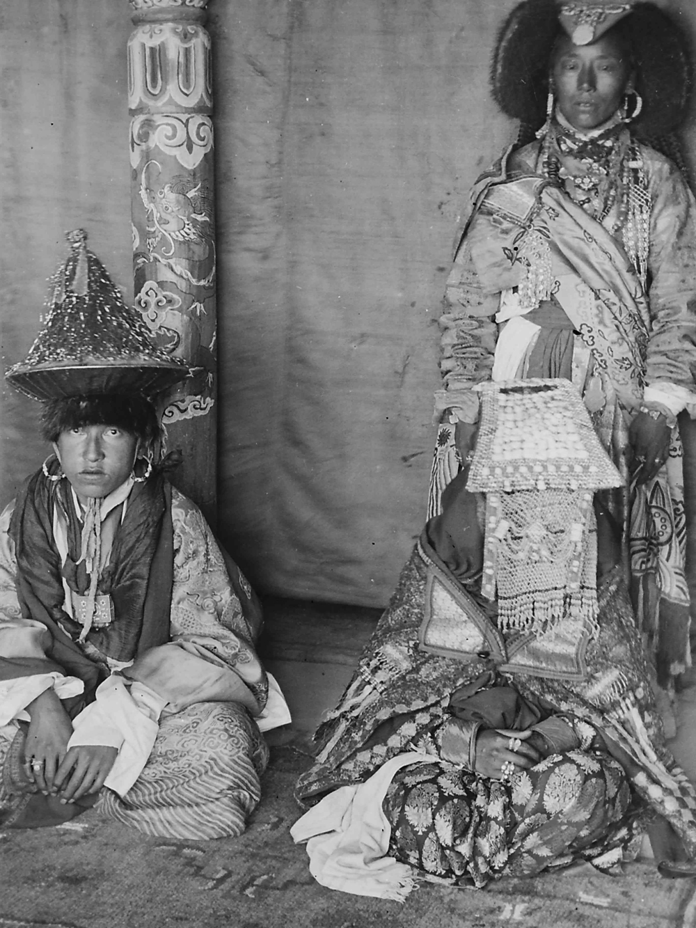 At the royal wedding in Stok, the crown prince Jigmet Dadul Namgyal and his bride queen Nyilza Wangmo are dressed in all their finery. The bride is dressed in an elaborate perag with a veil of basra pearls covering her face. Her perag is larger in the front than most typical ones worn by women in central Ladakh. She is also wearing a number of silver and gold rings. The crown prince is wearing a hat (serthop) covered with golden coloured threads, he wears large hooped earrings and around his neck hangs an amulet box (gau). (Photographer: Sebastian Schmitt, 1907–13; Courtesy: Gisela Müller)