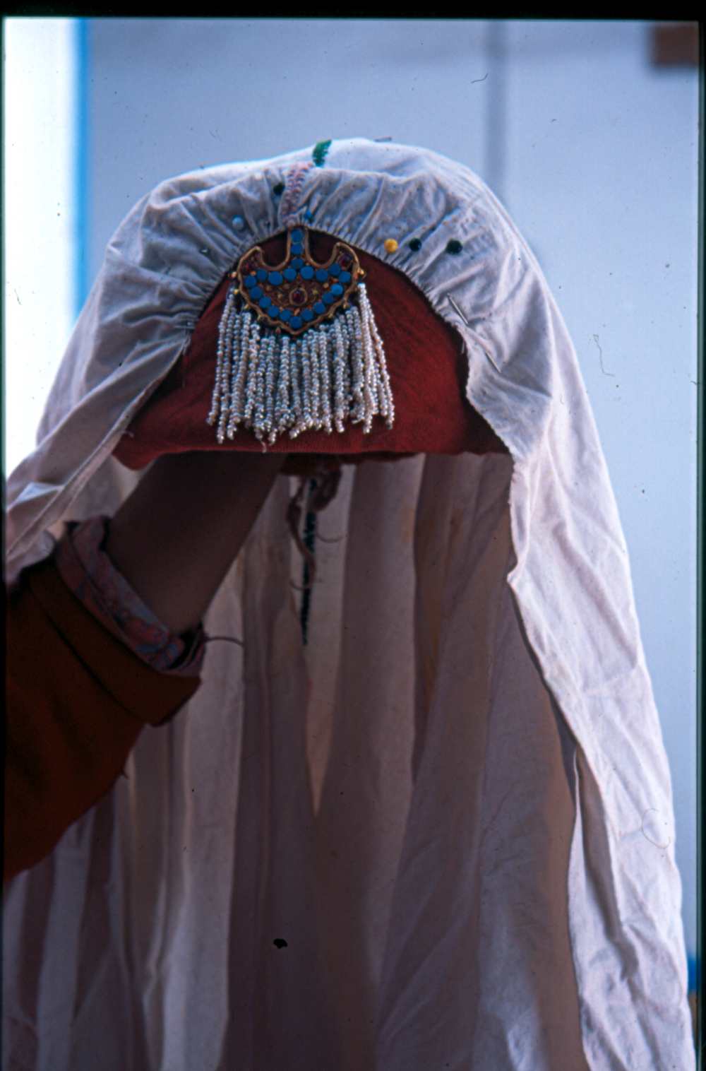 When Muslim women stopped wearing the perag, some say as late as the 1980s, they replaced it with the jhugni. This was a head ornament similar to the Indian jhumar, that is also worn in the centre of the forehead. The jhugni shown here is a half-crescent-shaped gold piece, with a central carnelian stone surrounded by turquoise. From the base of the crescent shape hang several strings of pearls. At the time of a wedding, the bride wears the jhugni along with a white cotton veil as shown here, this covers a woman’s head and extends halfway down her back. (Photographer: Monisha Ahmed, 2005)