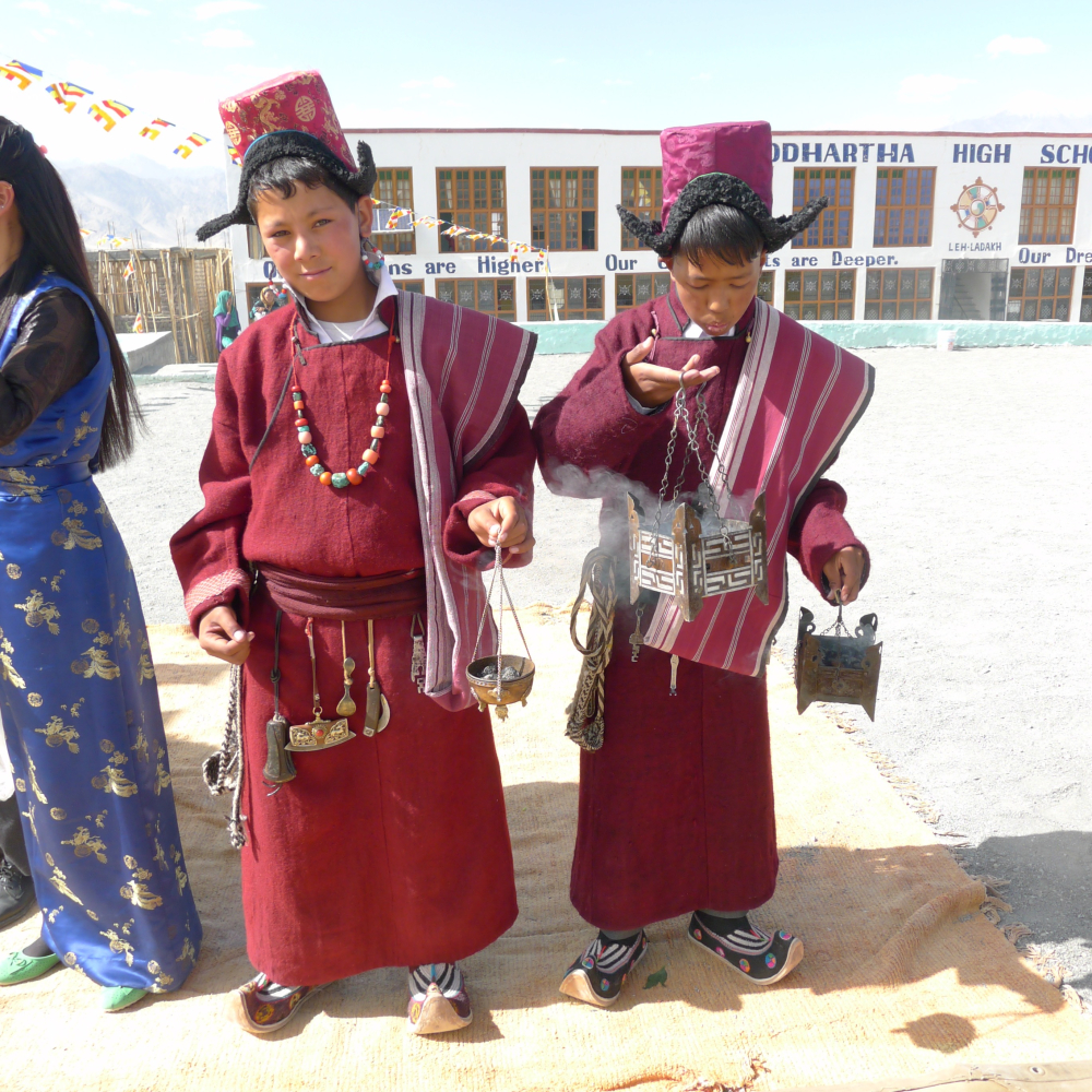 At a school ceremony, these young boys attend dressed in male Ladakhi attire. They are wearing woollen robes and silk brocade hats. From the belt of the boy on the left hangs, from left to right: a sling shot, leather needle case, metal ornamented flint purse, brass spoon, wood and metal knife and brass key. Around his neck is a necklace with coral and turquoise stones. In his ears are a loop of turquoise, imitation coral and pearl beads. (Photographer: Monisha Ahmed, 2015)